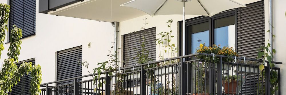 Moderner Balkon mit Sonnenschirm, Pflanzen und dunklen Jalousien.